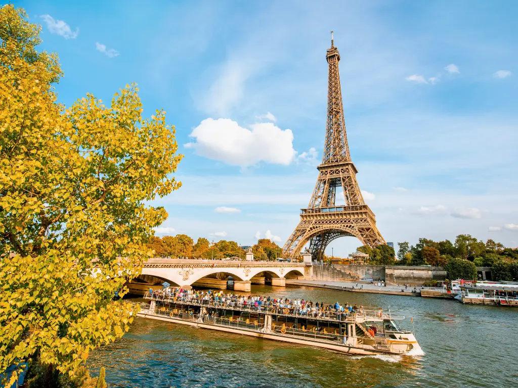 Autumnal hues of yellow leaves frame a Seine River cruise boat filled with passengers, with the Eiffel Tower and Pont d'Iéna bridge in the background under a soft blue sky with wispy clouds.