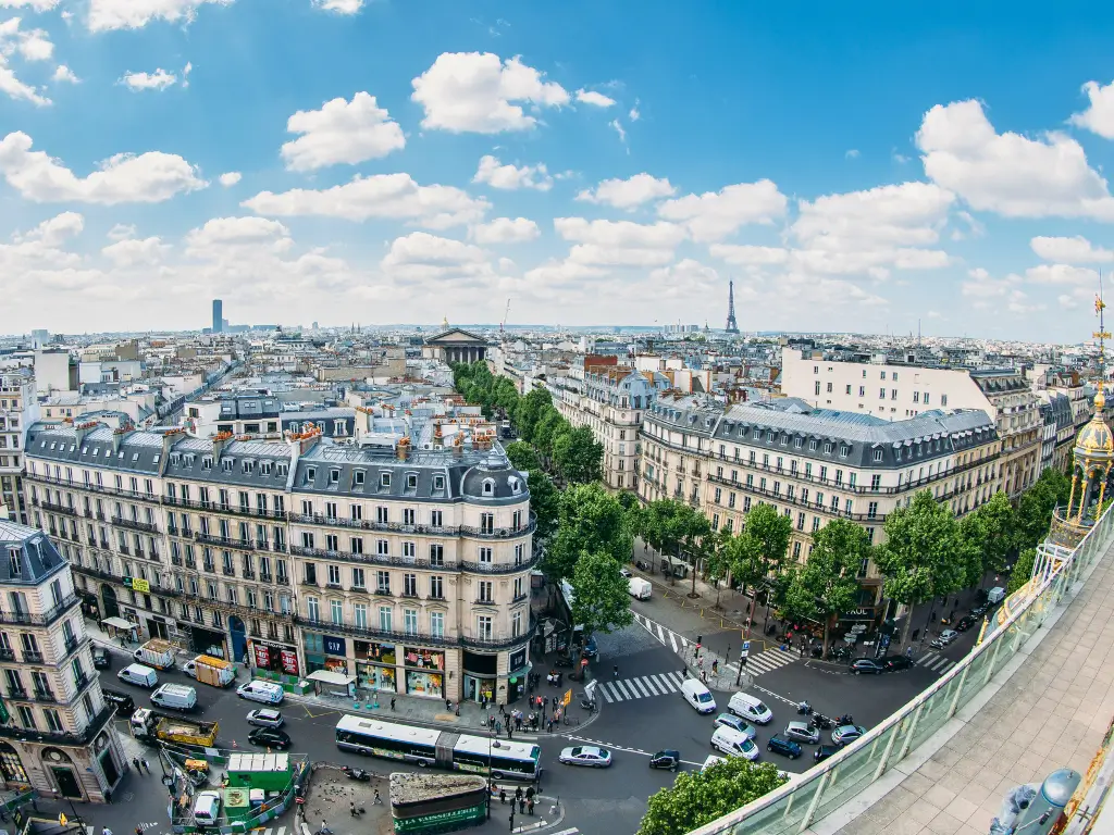 Panoramic view of Paris from Galeries Lafayette rooftop, showcasing the intricate Haussmannian architecture, bustling city streets with cars and pedestrians, and distant landmarks like the Eiffel Tower under a clear blue sky.