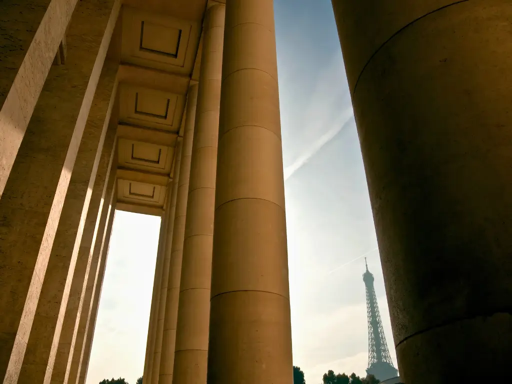 The Eiffel Tower peeking through the colossal stone columns of the Palais de Tokyo, creating a dramatic contrast between the modern monument and the classical architecture, with the warm glow of the sunset in the background.