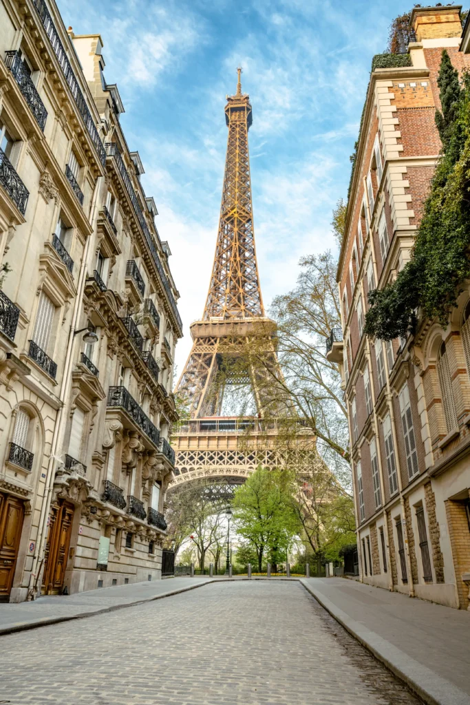 The Eiffel Tower looms large at the end of Rue de l'Université, flanked by classic Parisian buildings with ornate balconies, on a cobblestone street lined with budding green trees, capturing the essence of a Paris spring.