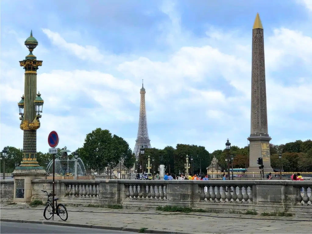 The Place de la Concorde in Paris, with its ornate lamp post and the Luxor Obelisk, the Eiffel Tower in the background, and a lone bicycle in the foreground against a cloudy sky.