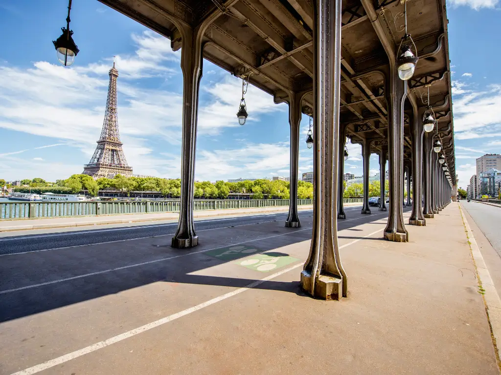 View of the Eiffel Tower from under the metal arches of Pont de Bir-Hakeim, featuring a series of ornate lampposts, on a bright sunny day with clear blue skies.