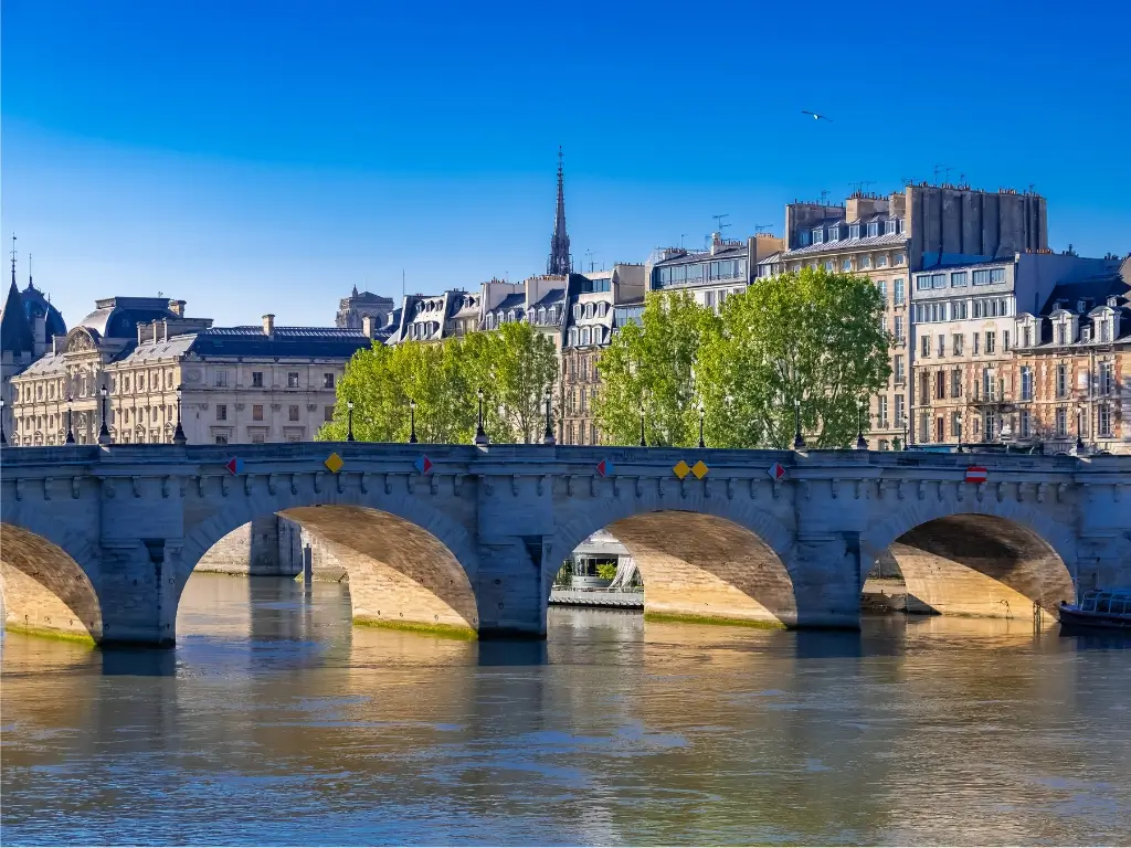 The Pont Neuf bridge crossing the Seine River with the Île de la Cité and its traditional Parisian buildings in the background, under a clear blue sky.