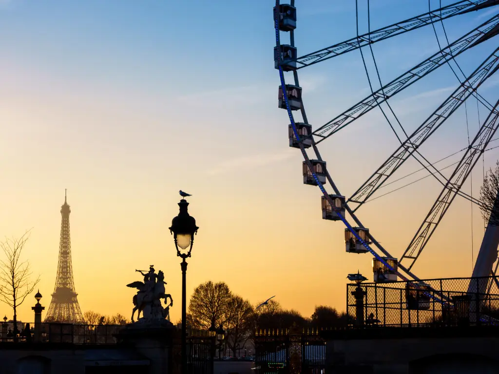 Silhouette of the Eiffel Tower at dusk with the Ferris wheel from Tuileries Garden on the right, and a classic Parisian street lamp adorned with a resting bird in the foreground, all against a golden sunset sky.