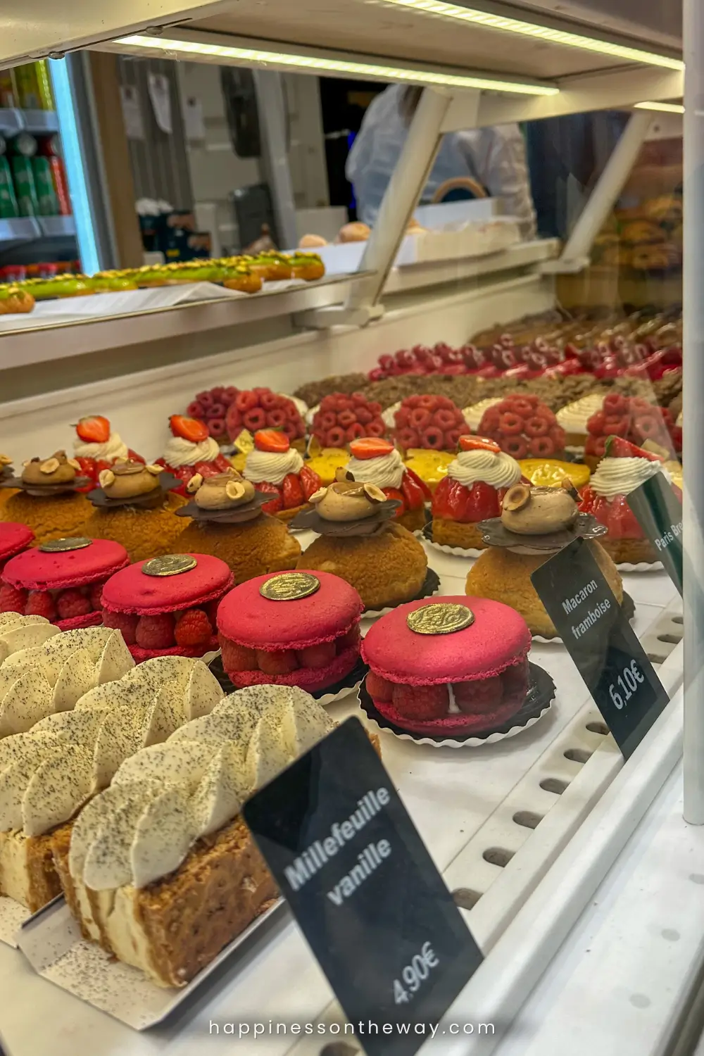 n enticing display of French pastries at a Parisian patisserie, featuring colorful macarons, fruit-topped tarts, and a classic millefeuille vanille. Price tags in the foreground suggest a customer's viewpoint, with the reflection of the bakery interior visible in the glass.