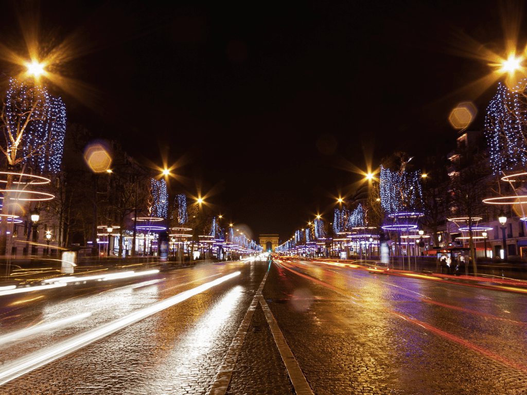 A vivid night scene on the Champs-Élysées, showcasing wet streets reflecting the glow of streetlights and illuminated trees adorned with blue Christmas lights, leading the eye towards a bright, vanishing point in the distance. This is why Paris is called the city of lights