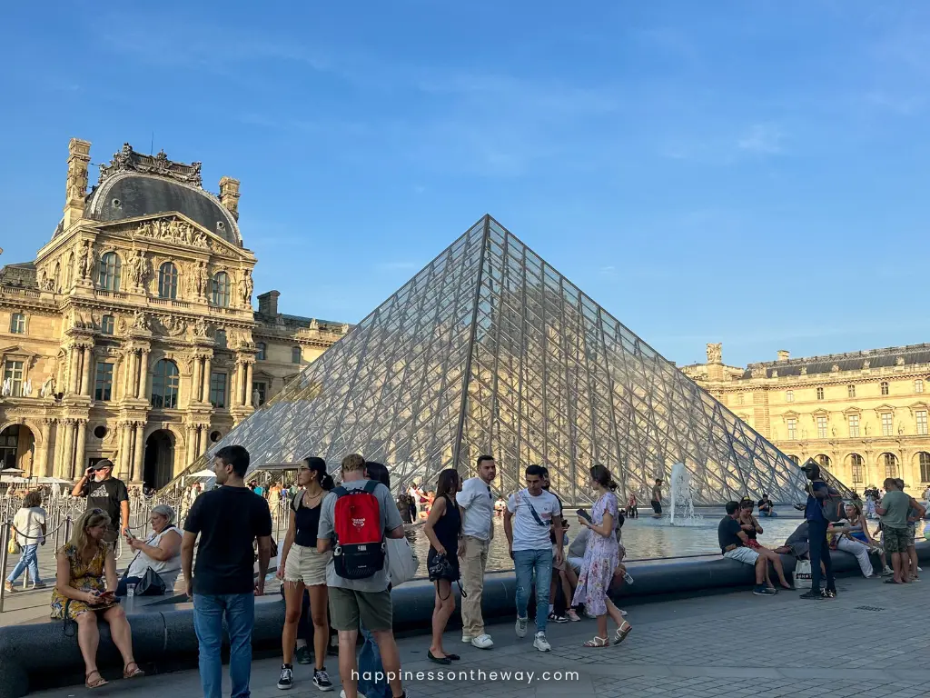 Tourists gather around the iconic Louvre Pyramid in front of the historical Louvre Palace under a clear blue sky in Paris. The contrast between the classical architecture and the modern glass pyramid creates a unique and photogenic landmark.
