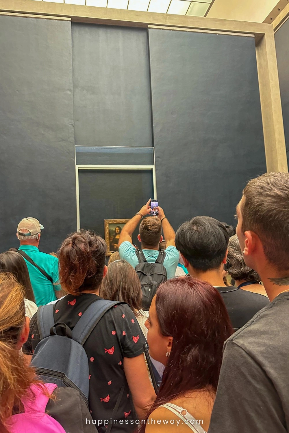 Is Paris overrated? A crowd of visitors with their attention fixed on the Mona Lisa, a man in the center raises his phone to capture a photo of the famous painting.