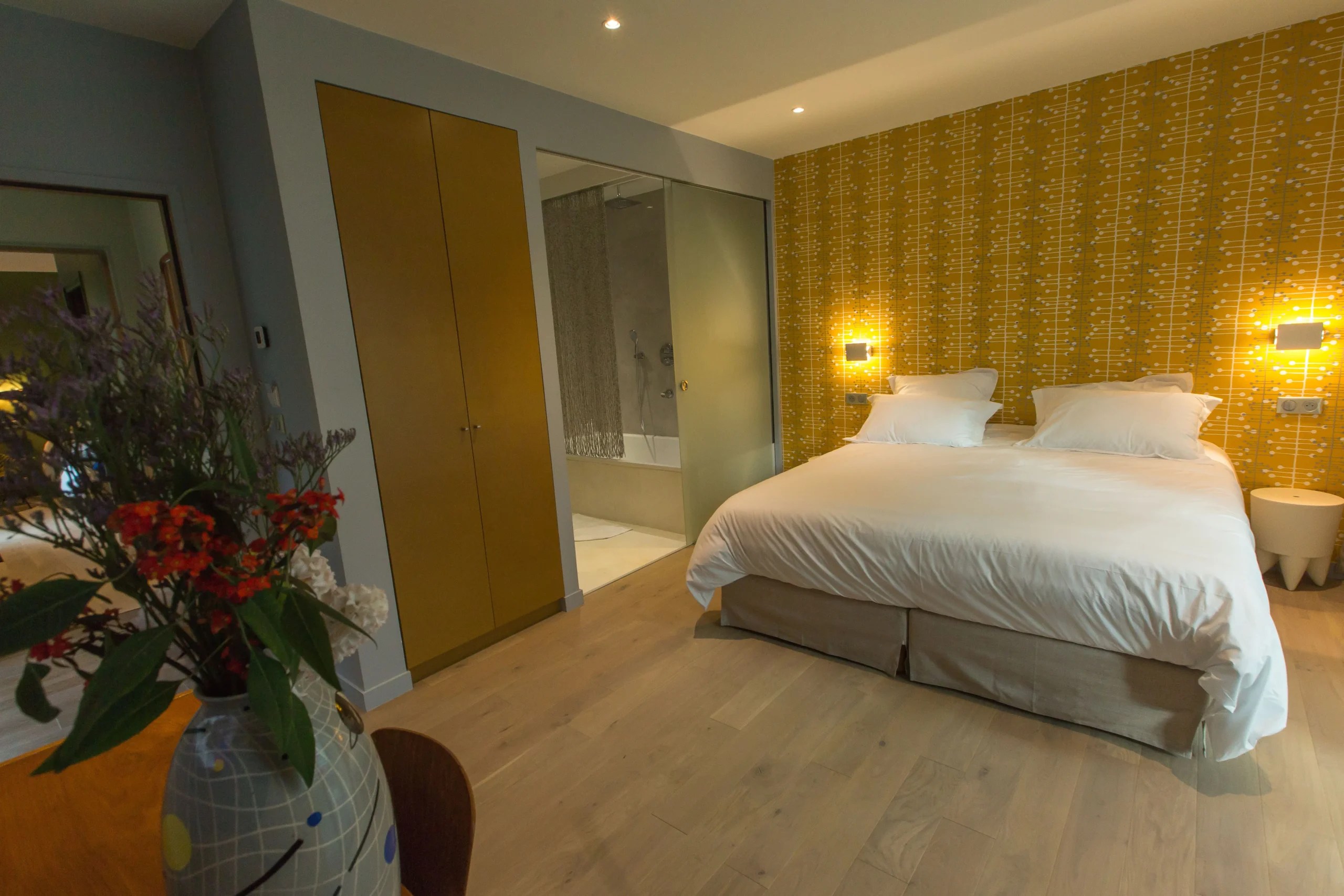Warm and modern room at Eden Lodge Paris, an eco-friendly hotel, with a cozy bed, yellow patterned wallpaper, and natural wood elements paired with a fresh bouquet.