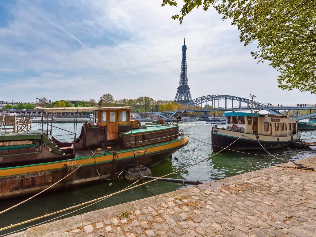 A view from the banks of the Seine River, with houseboats moored at the edge and the Eiffel Tower rising in the background, framed by greenery and a classic Parisian blue bridge, under a partly cloudy sky.