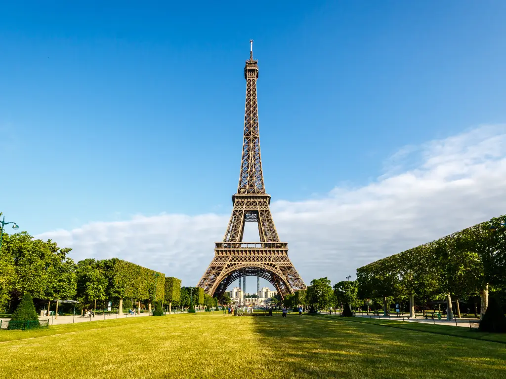 The Eiffel Tower stands majestically under a clear blue sky, centered in the frame from the Champ de Mars. The well-maintained green lawns are famous spots for Eiffel Tower picnic.