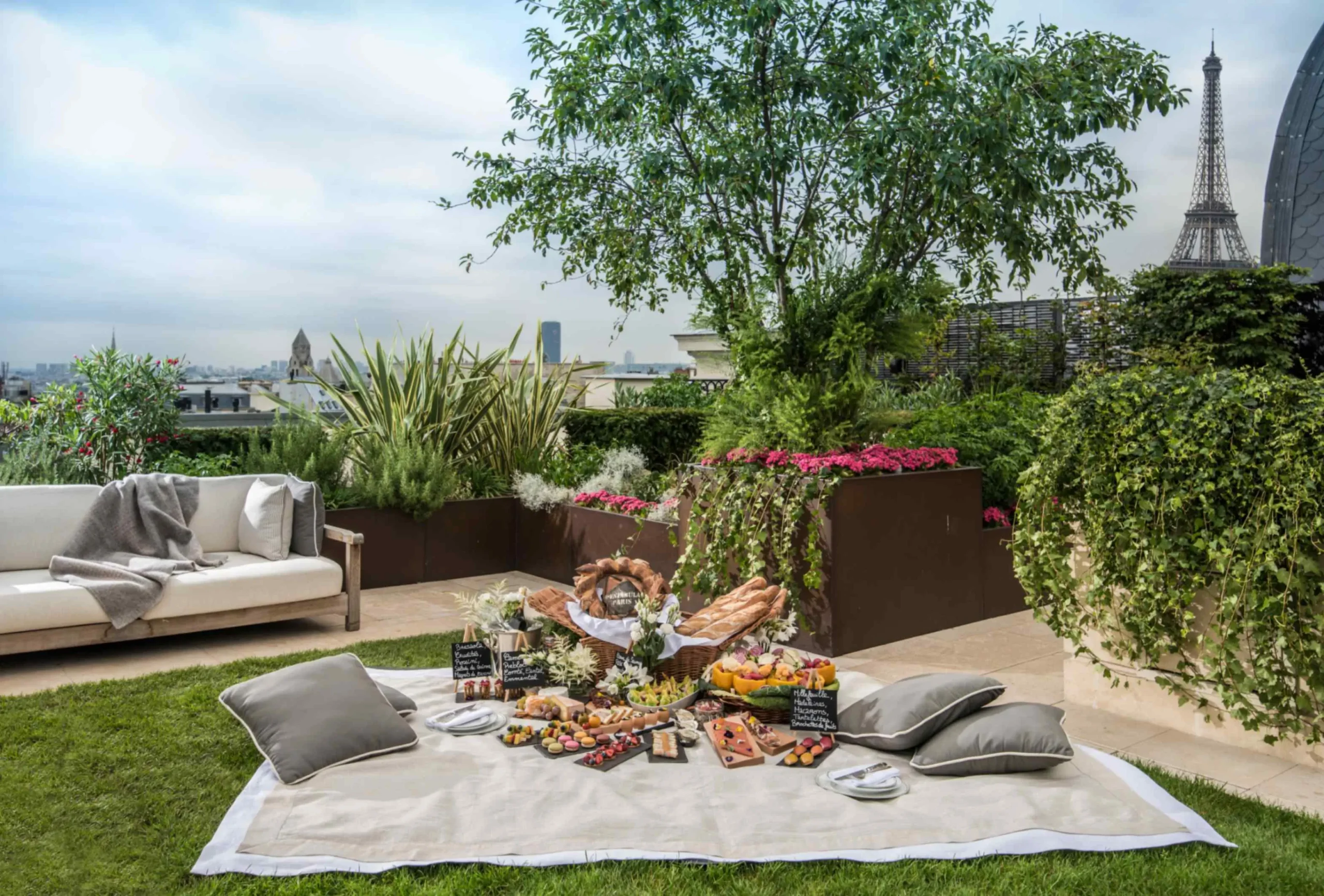 Luxurious rooftop picnic setup at Peninsula Paris, an eco-friendly hotel in Paris, with a spread of gourmet snacks on a blanket amidst lush garden greenery, offering a stunning view of the Eiffel Tower in the background.