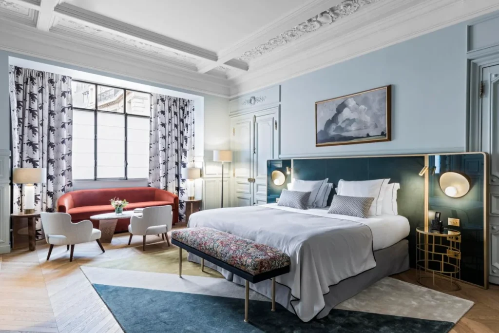 Stylish and sustainable guest room at Grand Powers Paris, featuring a comfortable bed with grey and white bedding, a red sofa, modern art, and a harmonious blend of classic and contemporary design elements.