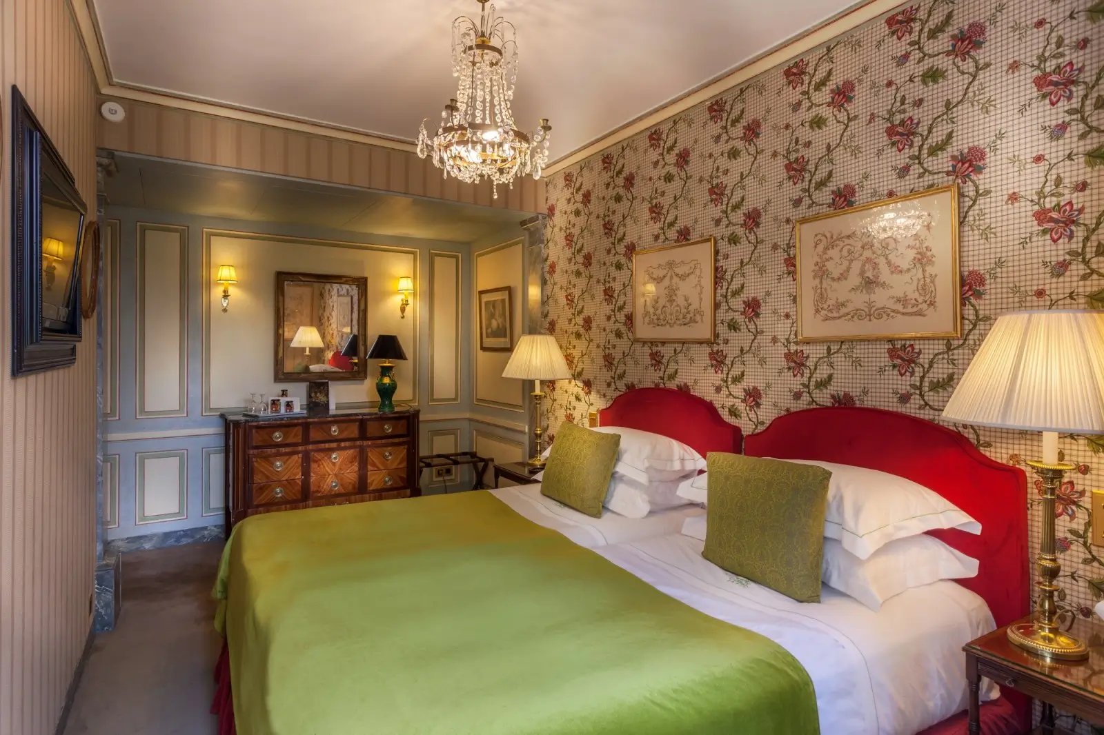 Classic Parisian charm in an eco-friendly setting at Hotel Duc de Saint Simon, featuring a luxurious bedroom with vintage floral wallpaper, a chandelier, and plush red and green bedding.