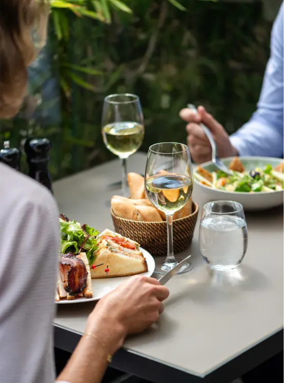 Intimate dining experience at Hotel Napoléon's eco-friendly bar in Paris, showcasing a meal of fresh salad and quiche, accompanied by white wine and a basket of bread, set against a backdrop of greenery.