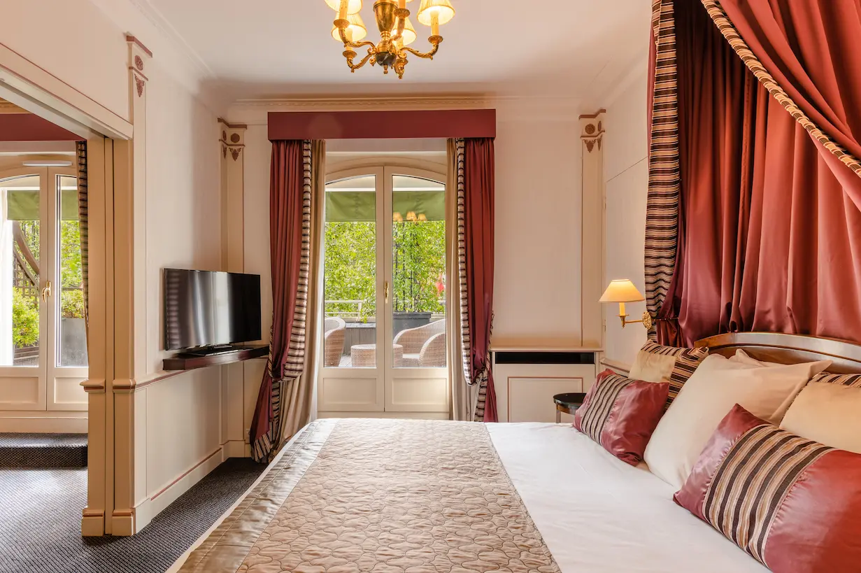 Classic Parisian charm meets sustainability in Hotel Napoléon's guest room, featuring a plush bed with striped and maroon accents, ornate curtains, and a balcony view of green foliage, aligning with eco-friendly hotel standards.