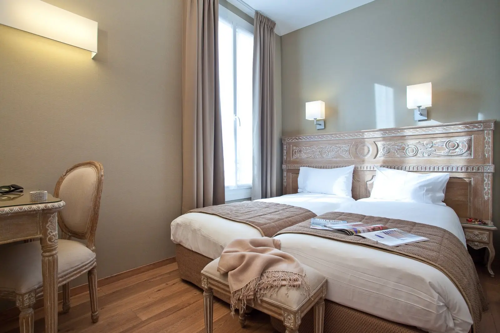Cozy and sustainable bedroom at Hotel du Printemps, an eco-friendly hotel in Paris, featuring a traditional carved wooden bed frame, soft beige tones, and a quaint sitting area by the window with views of the city.