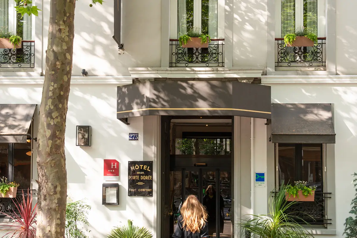 Entrance of Hôtel de la Porte Dorée in Paris, nestled among trees, with a woman entering the environmentally friendly establishment, which boasts a serene facade with balcony planters.