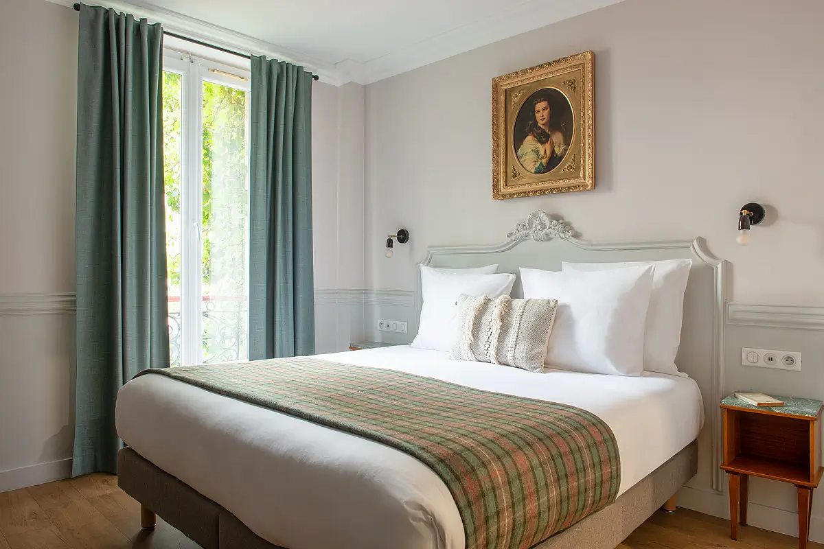 Classic French elegance with an eco-conscious twist at Hôtel de la Porte Dorée, showcasing a comfortable bed with a green striped throw and a vintage portrait in a gilded frame.