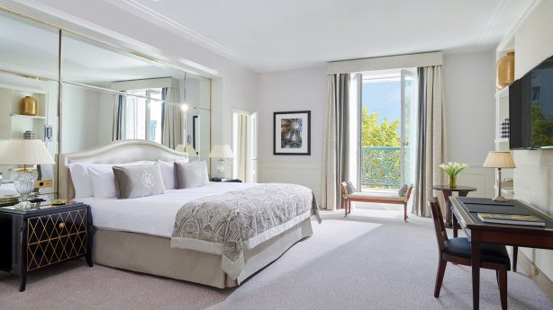 Luxurious suite at InterContinental Paris Le Grand, featuring a sumptuous bed with decorative throw, a well-appointed desk area, and French doors leading to a balcony with greenery, offering a serene urban oasis.