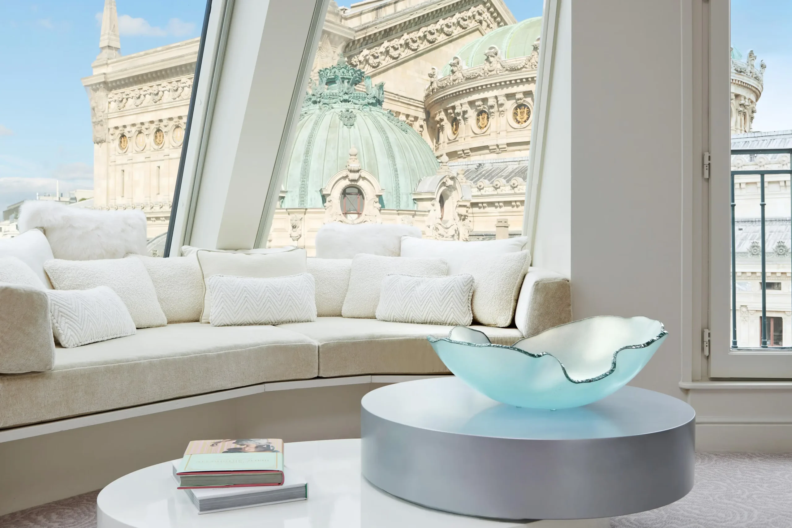Luxurious and sustainable lounge area at the Intercontinental Paris, with plush white cushions, an elegant aqua glass bowl on a coffee table, and a grand view of the Palais Garnier opera house.