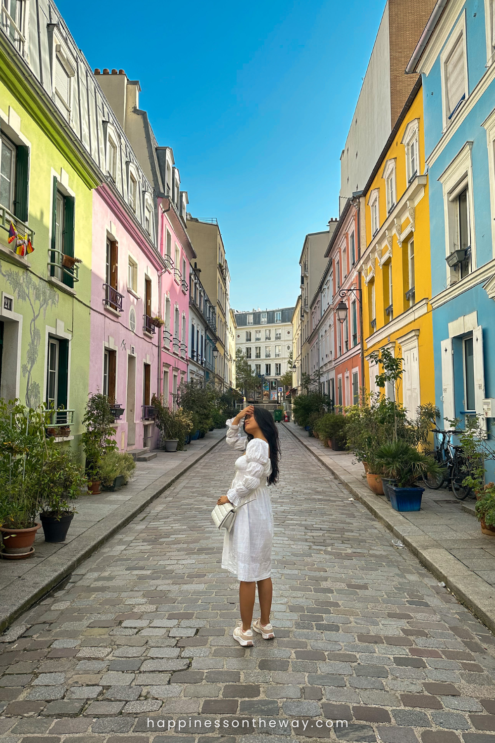 Me in a white dress stands on a cobbled street, her hand gracefully touching her hat, with the colorful facades of Rue Crémieux in the background.