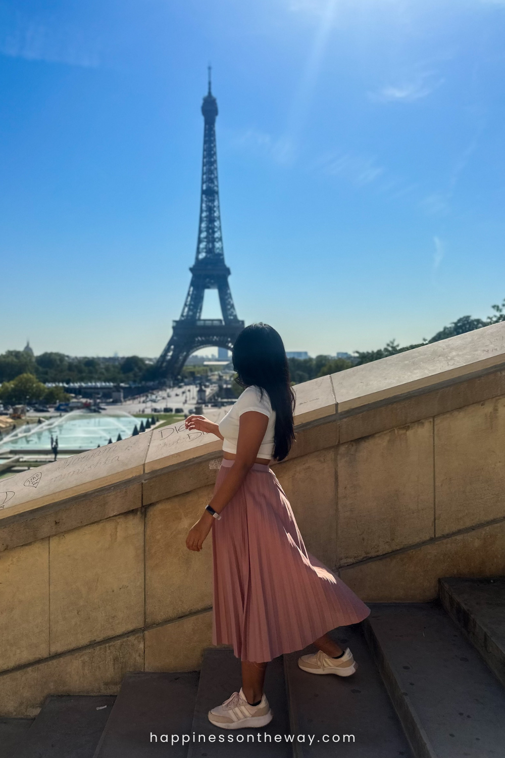 Me in in a pink pleated skirt and white top gazing at the Eiffel Tower, walking on steps on a sunny day. A questioning concept emerges, 'Is Paris overrated?'