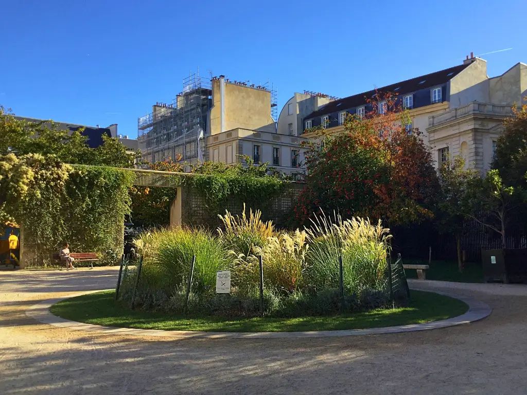 A peaceful scene at Jardin Anne-Frank, one of the best picnic spots in Paris. It shows a rustic pergola casting shadows over a circular bench, surrounded by tall grasses and urban greenery, contrasting with the Parisian buildings in the background.