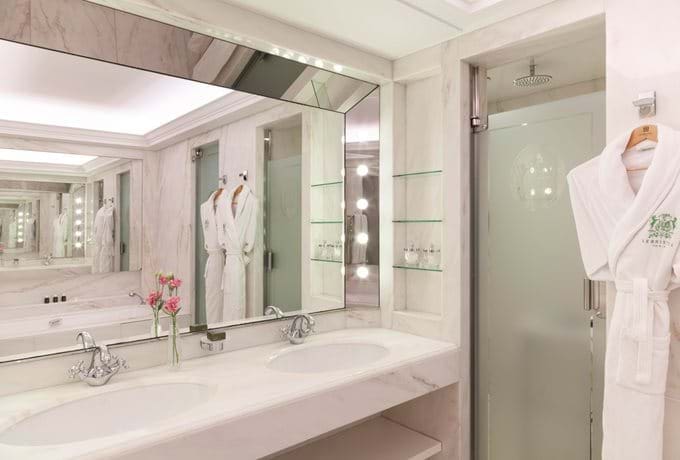 Elegant and bright bathroom at Le Bristol Paris, featuring dual sinks with marble countertops, mirrored walls, and plush white robes, highlighting the hotel's commitment to eco-friendly opulence.