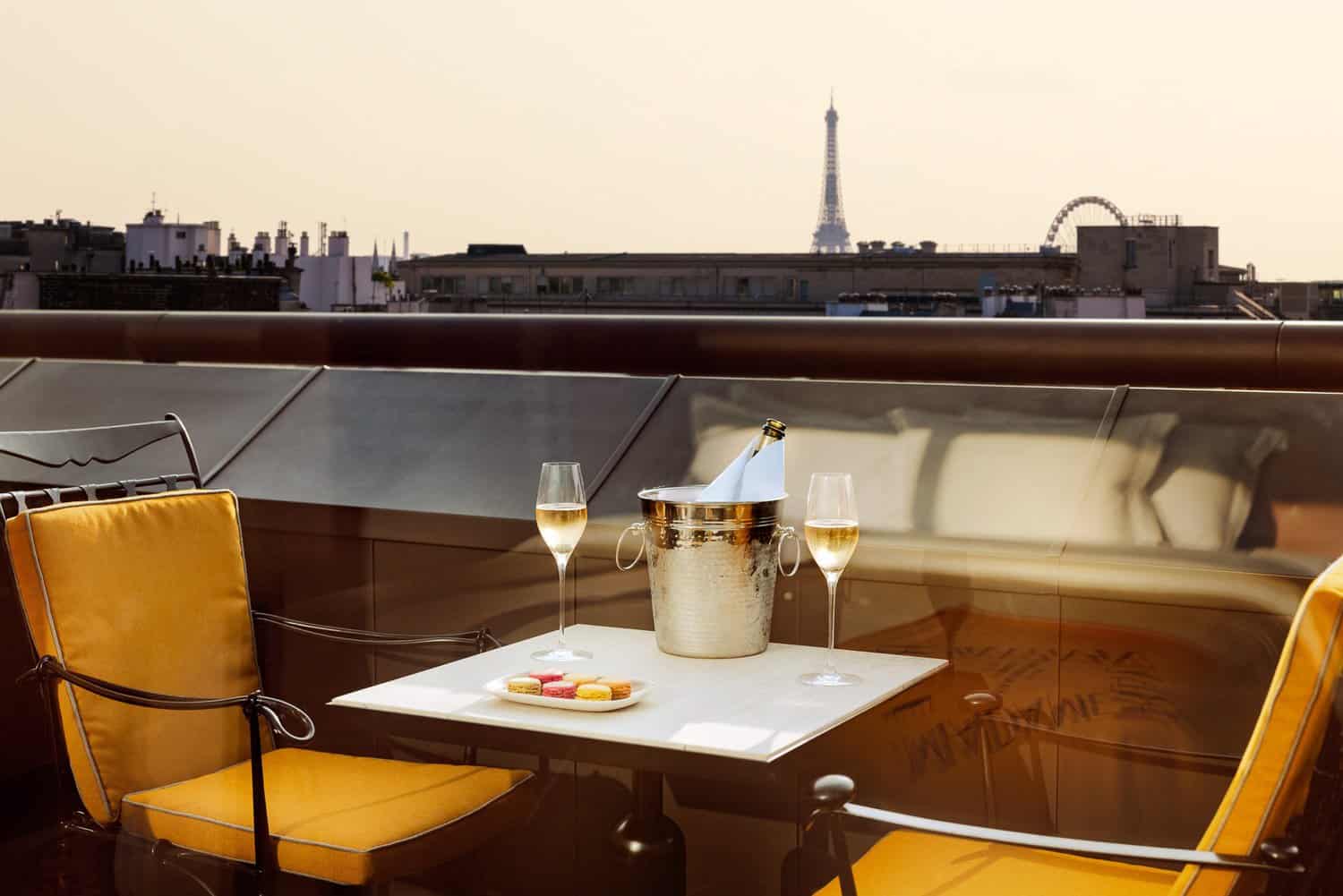 Luxurious relaxation on the Madame Rêve Hotel terrace with a close-up of champagne in a silver bucket, two glasses, and French macarons, overlooking the Eiffel Tower at sunset, symbolizing eco-luxury in Paris.