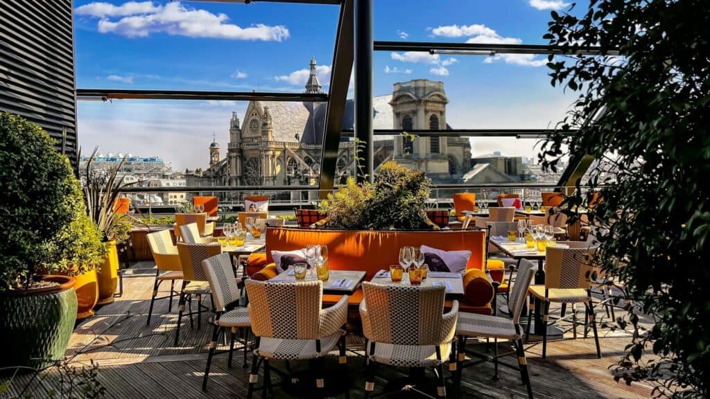 Elegant rooftop dining setup at Madame Rêve Hotel in Paris, featuring orange cushions and wicker chairs, with a view of historical buildings under a blue sky, conveying an eco-friendly hotel experience.