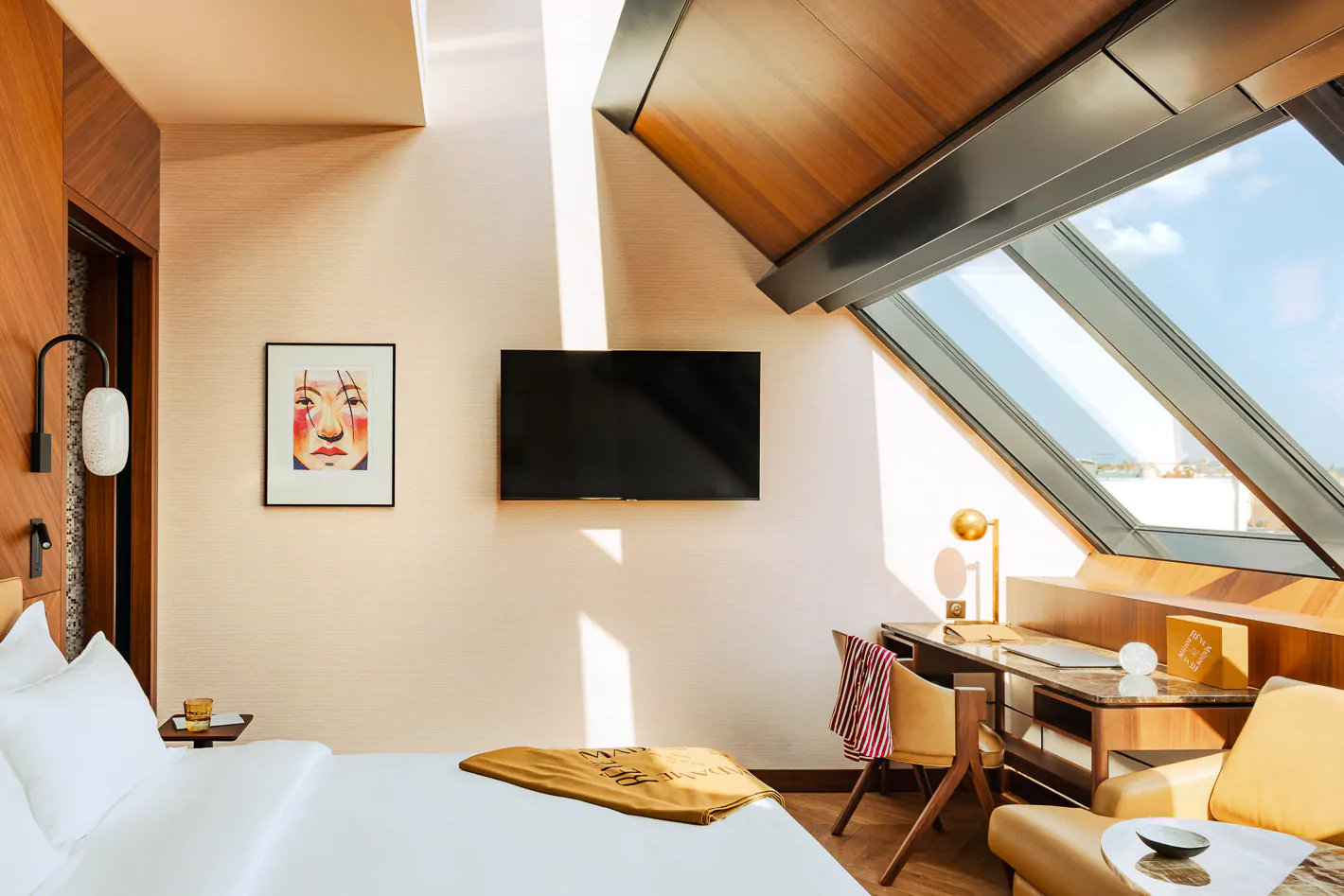 Chic and modern room at Madame Reve in Paris, with a sleek design featuring a plush bed, striking artwork, and a wall-mounted TV. Natural light pours in through the large angled window, offering a glimpse of the Parisian skyline.