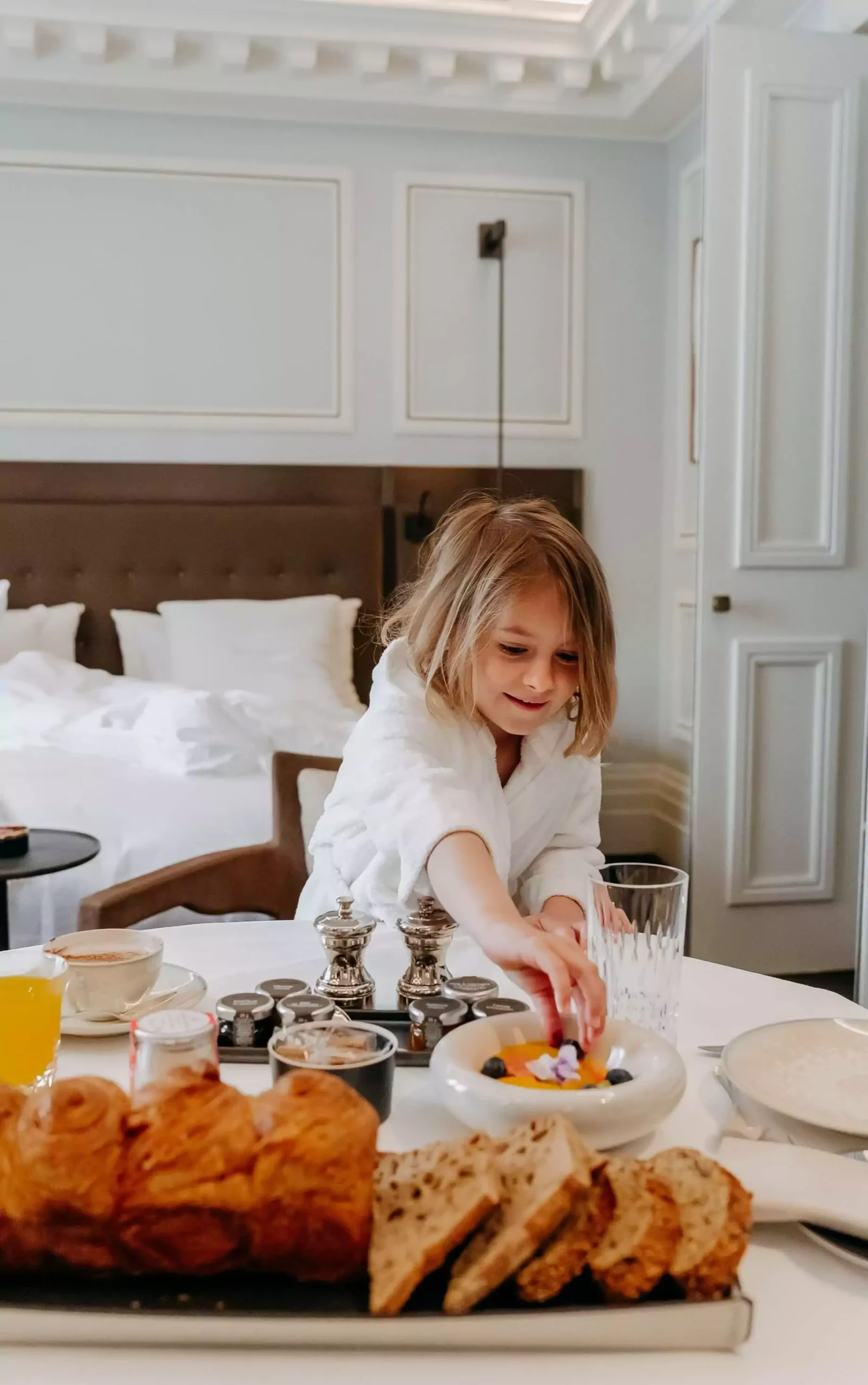 A young child in a white bathrobe reaching for berries from a healthy breakfast spread in a luxurious room at Maison Villeroy, an eco-friendly hotel in Paris, with a cozy bed and elegant decor in the background.