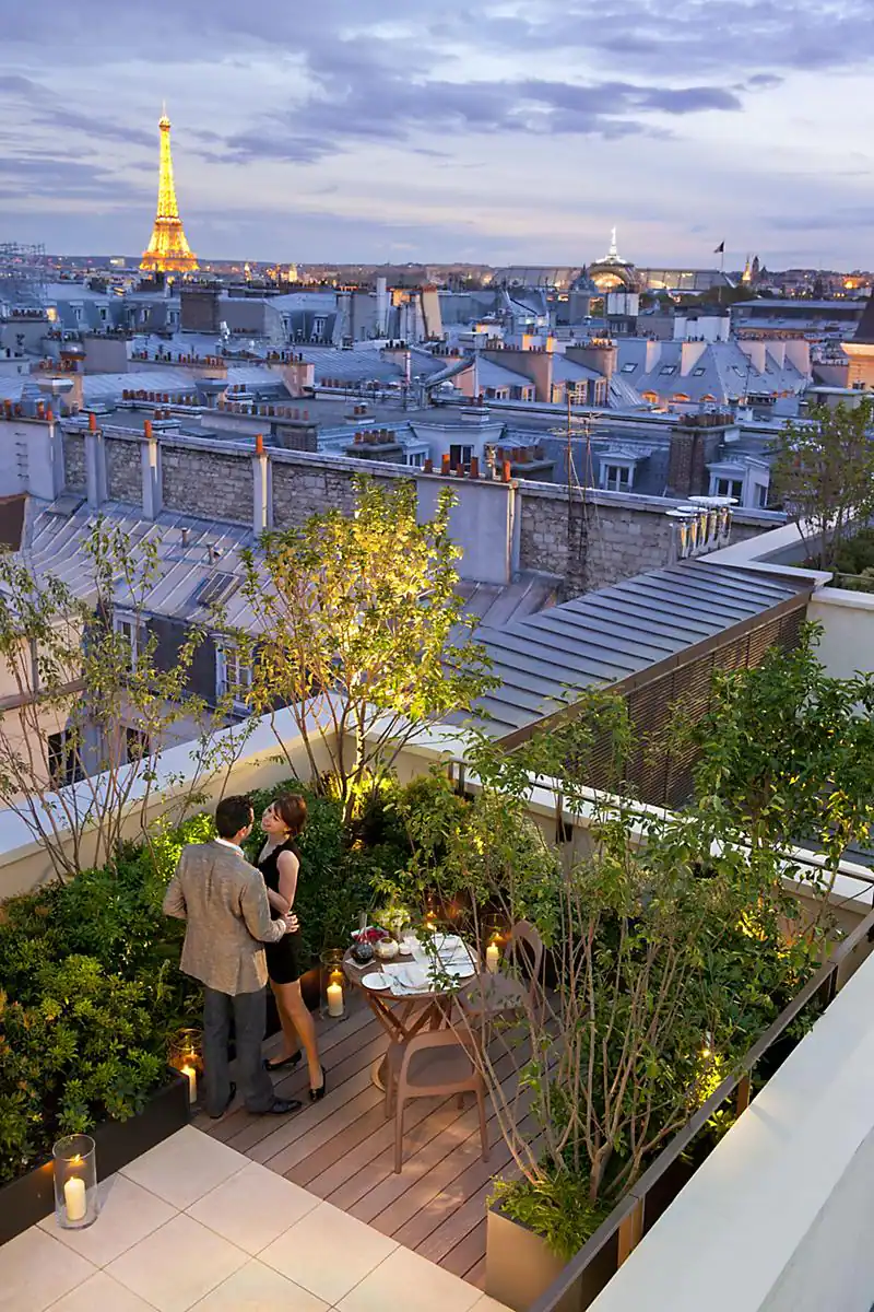 Romantic rooftop setting at the Mandarin Oriental, a sustainable hotel in Paris, with an intimate dining area surrounded by lush greenery and candles, offering a breathtaking view of the Eiffel Tower at twilight.