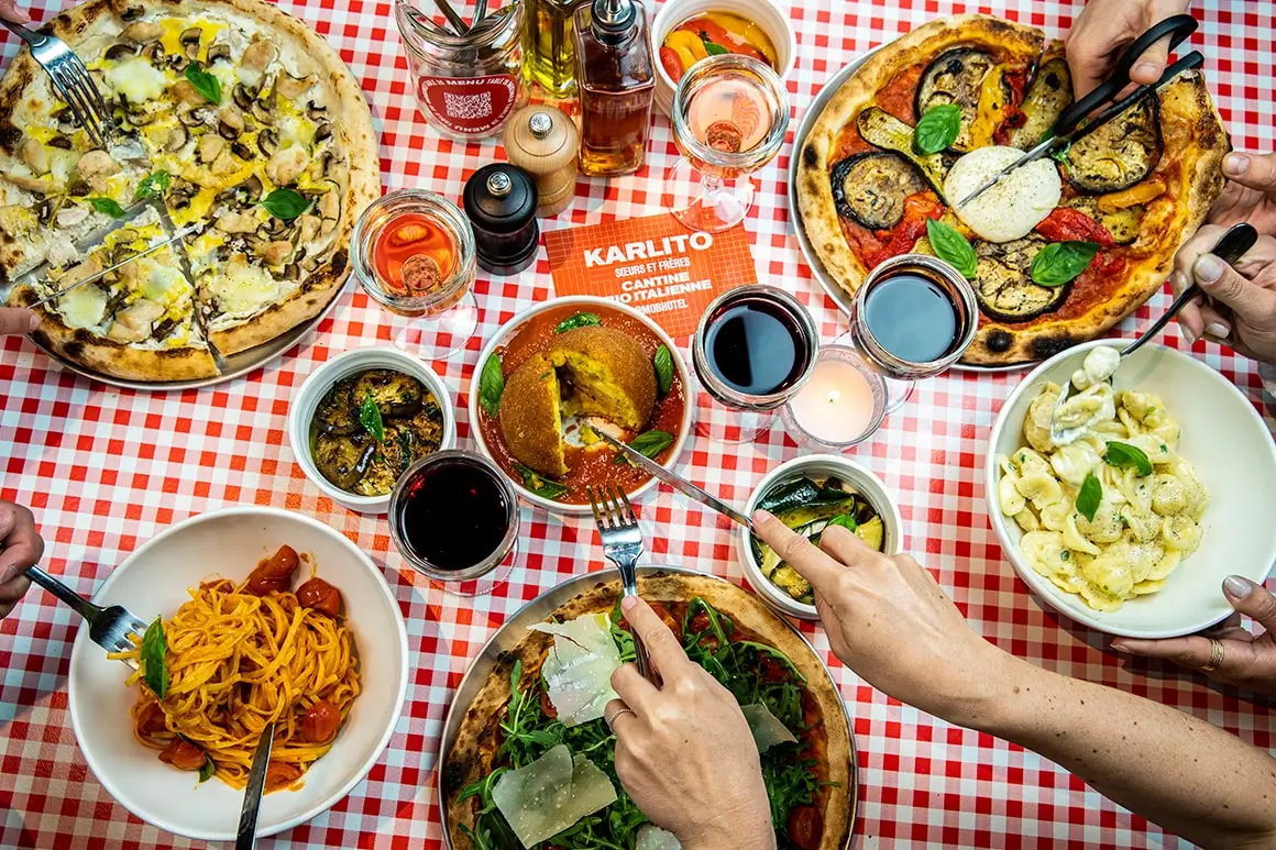 A vibrant feast at Mob Hotel's organic canteen in Paris, showcasing a variety of pizzas and pastas with colorful toppings, glasses of wine, and a casual communal dining atmosphere.