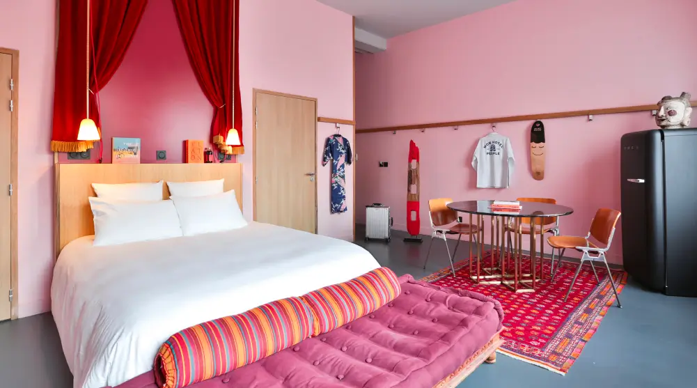Eclectic and colorful guest room at Mob Hotel in Paris, featuring a comfortable bed with bold textiles, pink walls, and quirky decor, reflecting the hotel's eco-conscious ethos.