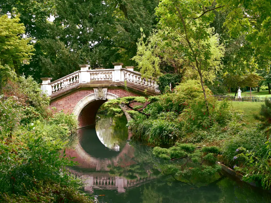 A romantic red brick and stone bridge arches gracefully over a calm stream in Parc Monceau, one of the best parks in Paris for picnic, surrounded by lush greenery and tranquil walking paths, evoking a serene escape within the city.