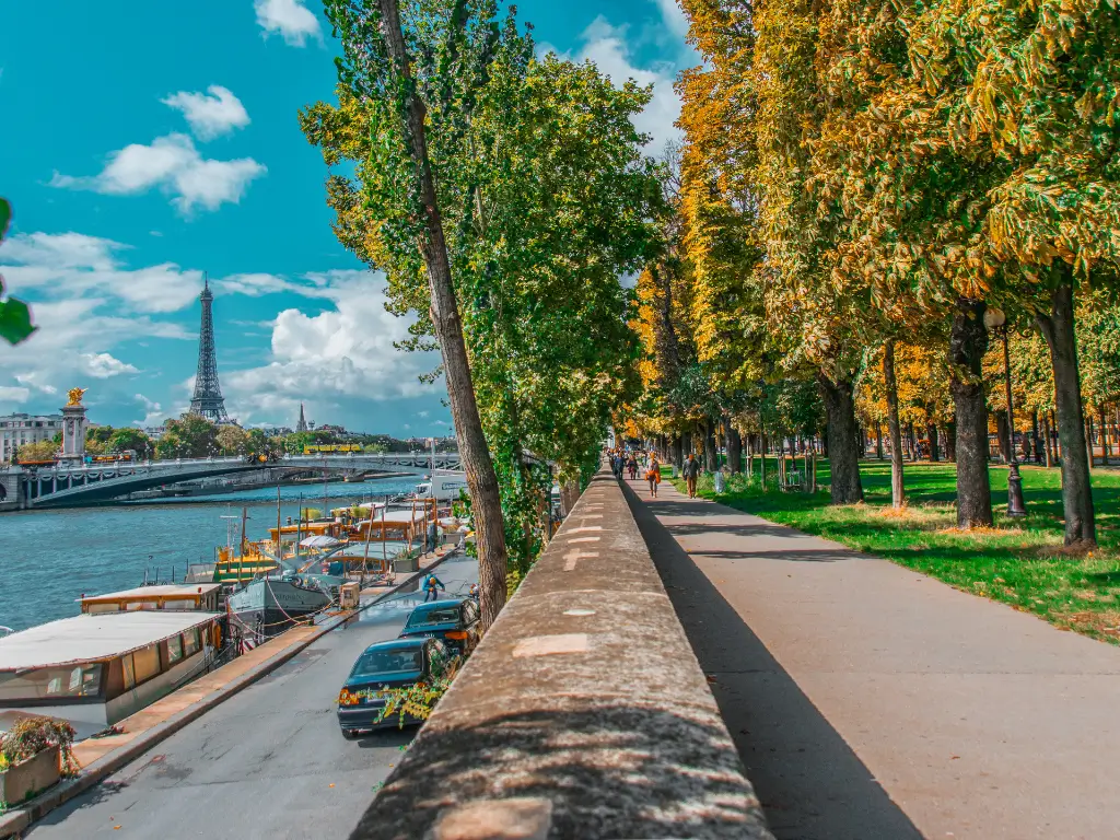 A picturesque riverside promenade at Parc Rives de Seine, a place in Paris for picnic. With golden autumn trees lining the path, views of the Eiffel Tower in the distance, and Parisians enjoying a leisurely day.