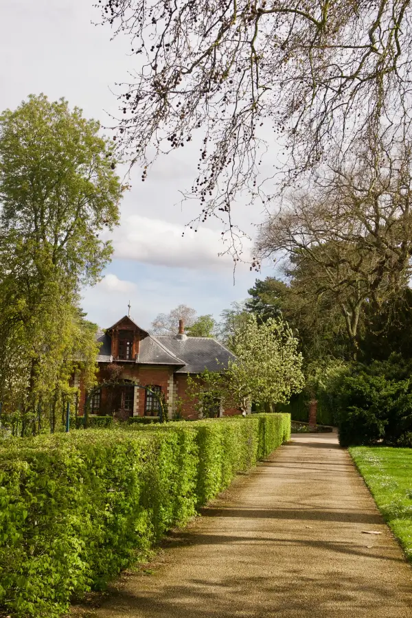 A quiet pathway in Parc de Bagatelle, one of the best picnic spots in Paris, leading to a charming red-brick house, framed by early spring foliage and a well-maintained hedge, evoking a sense of peaceful retreat.