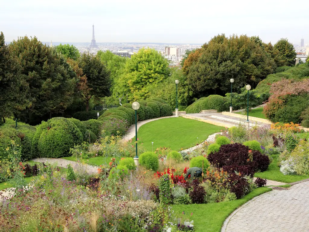 Parc de Belleville is one of the best parks in Paris to picnic with panoramic views of Paris, with the Eiffel Tower in the distance, amidst a diverse array of garden beds and shrubbery, neatly arranged on rolling hills and accompanied by paved pathways.