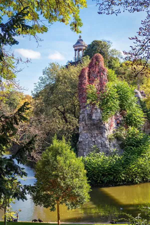 View of the Parc des Buttes-Chaumont with a picturesque stone cliff partially covered in red ivy, topped by a classical-style gazebo, alongside plants and trees and a pond reflecting the blue sky. This park is one of the best packs in Paris to picnic