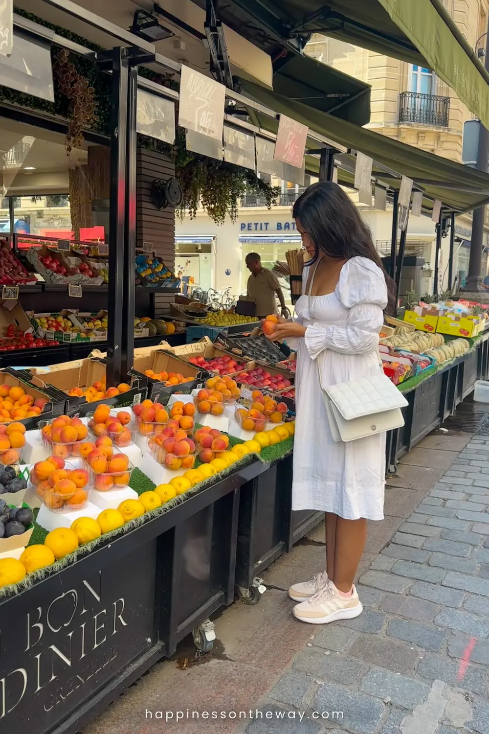 A woman in a white summer dress and sneakers is examining oranges at the Au Bon Jardinier fresh fruit stall at Rue Cler market in Paris. The market display is vibrant with assorted fruits, and the scene captures a typical sunny day with patrons browsing in the background, highlighting the local Parisian shopping experience.
