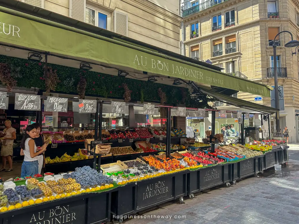 A bustling fruit stand, "Au Bon Jardinier," on Rue Cler in Paris, brimming with an array of fresh produce under a canopy of green awnings, with shoppers browsing and a woman in the foreground checking her phone.