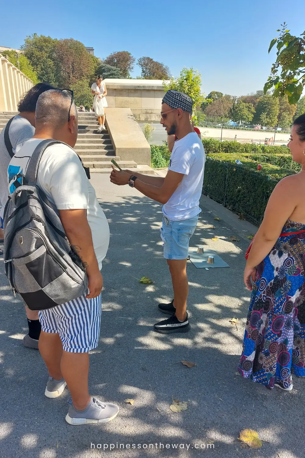 A man in a white shirt and patterned cap is engaging with onlookers in a classic ball and cups guessing game known as shell game or thimblerig scam on the Trocadéro esplanade in Paris.