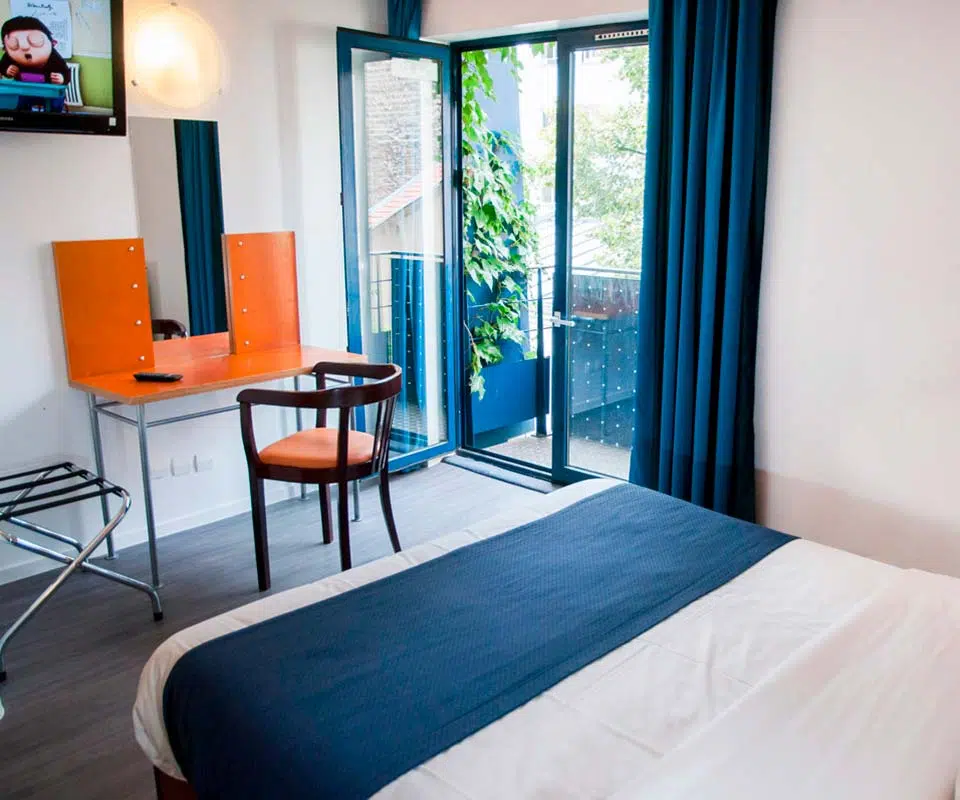 Bright and inviting eco-friendly guest room at Solar Hotel Paris, featuring a clean white bed with blue accents, large windows, and a city view, promoting sustainable travel.