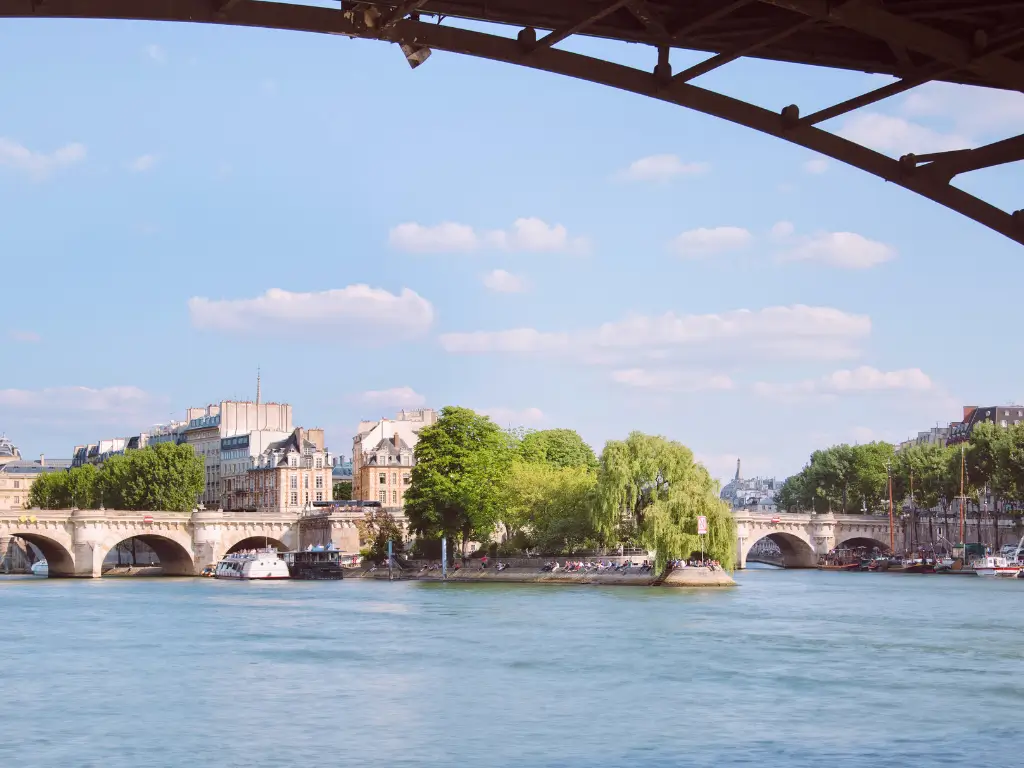 Underneath the arches of a Parisian bridge, the image captures the calm waters of the Seine River, a glimpse of the city's architecture, and the leafy Square du Vert Galant, one of the best parks in Paris to picnic, with the Eiffel Tower in the distance.