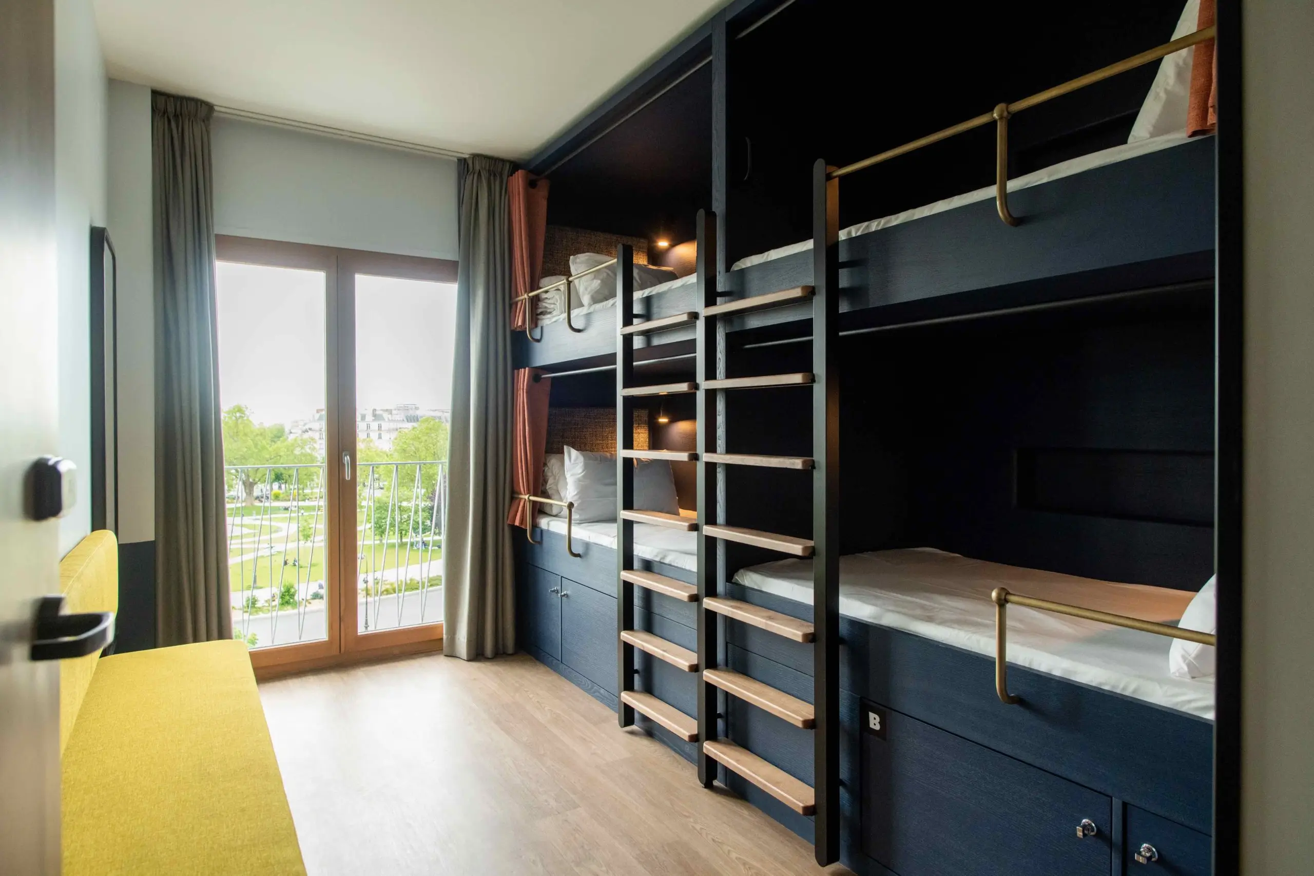 Modern and efficient dormitory room at The People Paris Nation hostel with sleek bunk beds, offering a view of the Parisian streetscape through a balcony door.