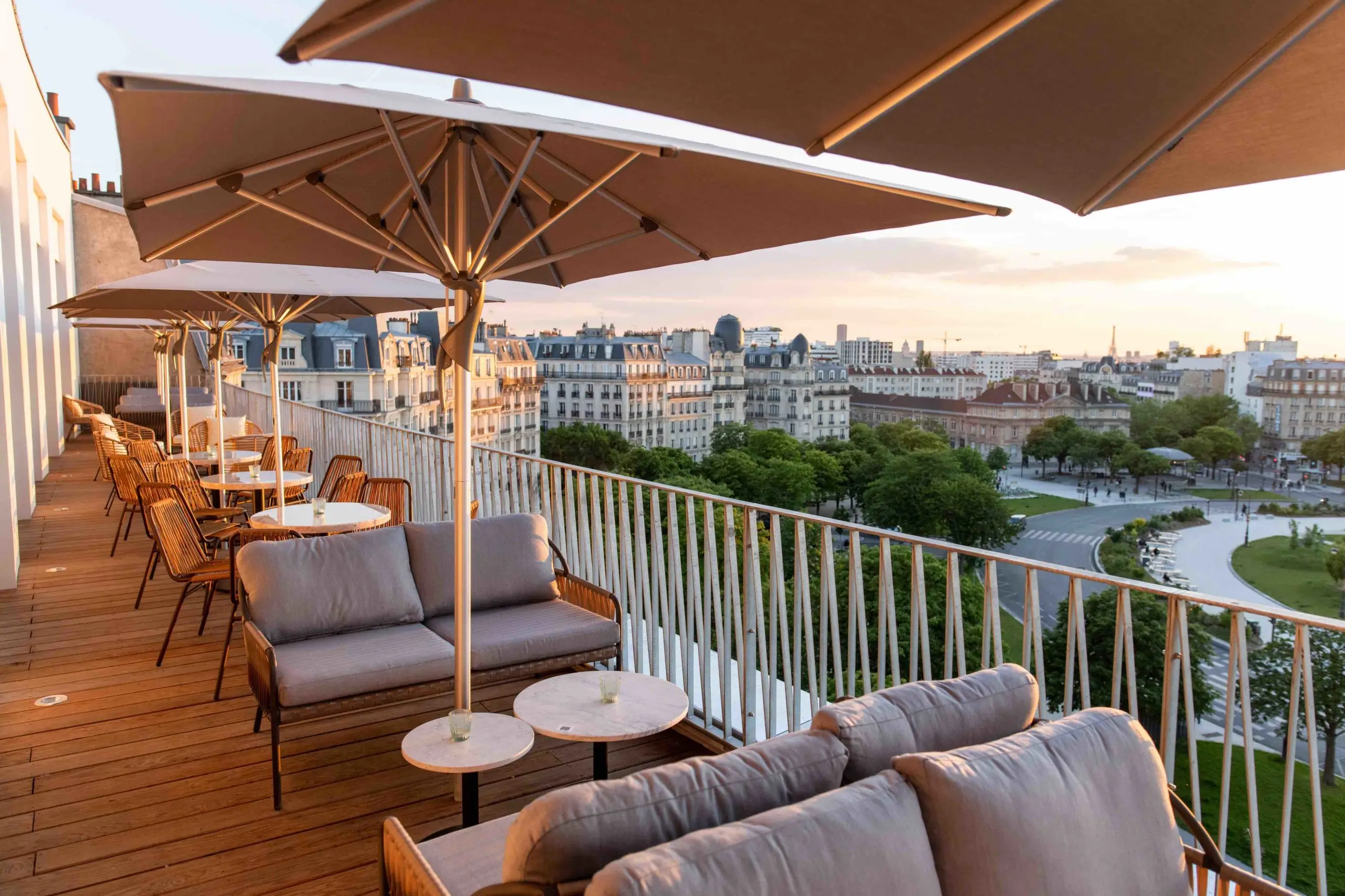 View from The People - Paris Nation, one of the eco-friendly hotels in Paris featuring a serene rooftop terrace with plush seating, wooden tables, and large umbrellas, overlooking a picturesque Parisian cityscape at dusk.
