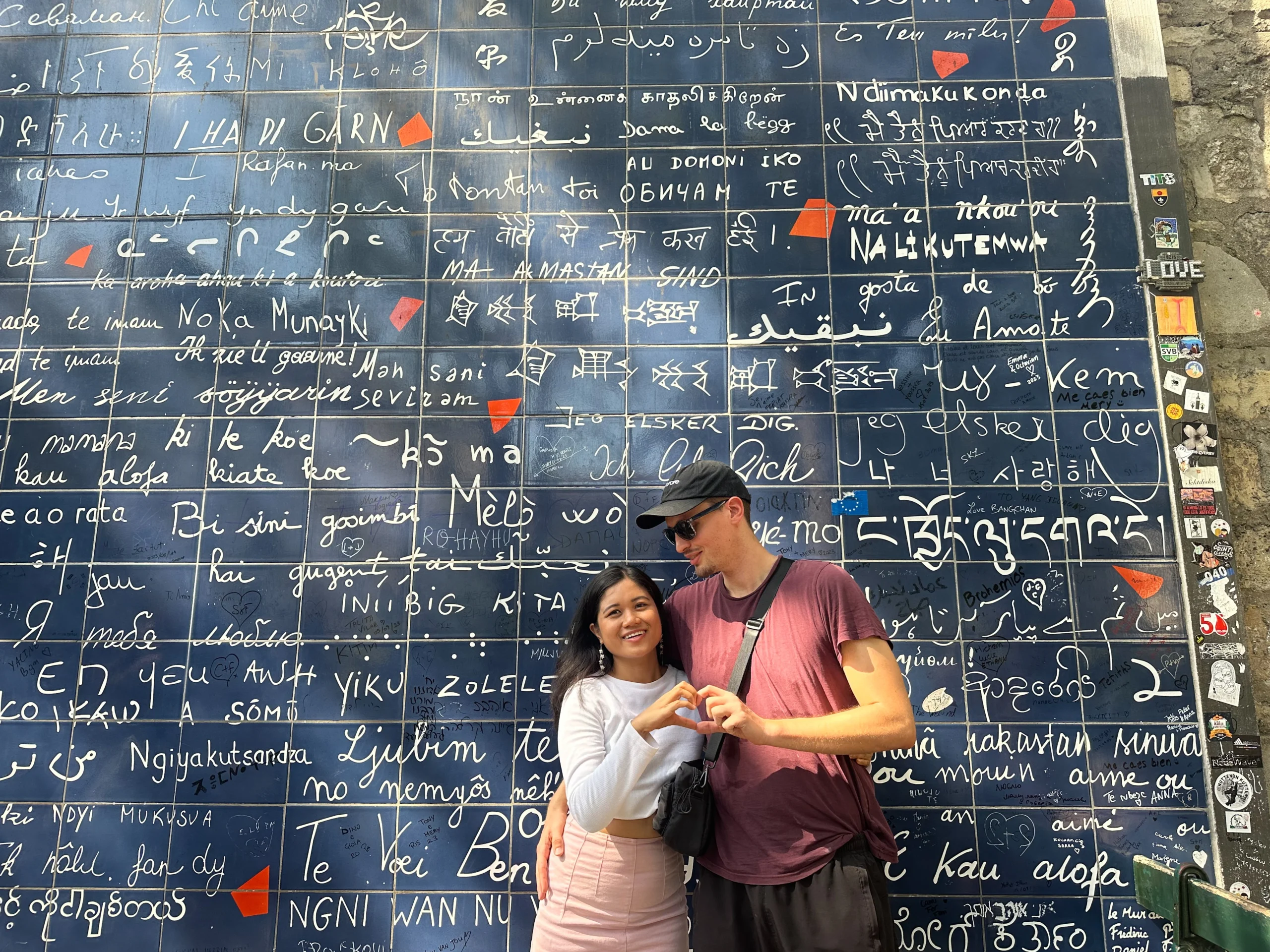 Me and my partner, Mat form a heart shape with their hands in front of the "Wall of Love" in Paris, where the phrase "I love you" is written in various languages on blue-tiled backdrop, a testament to the universal language of love.