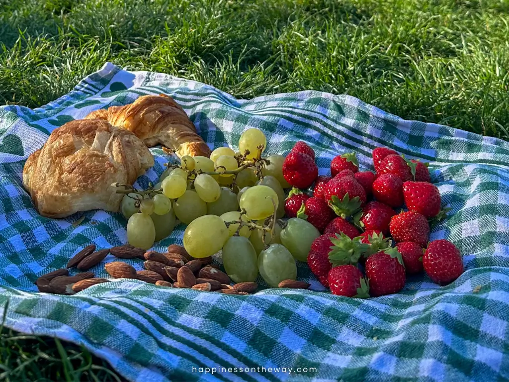 A close-up of a picnic spread on a blue and white checkered cloth featuring ripe strawberries, a bunch of green grapes, a flaky croissant, and scattered almonds, set on green grass for a Champ de Mars picnic.